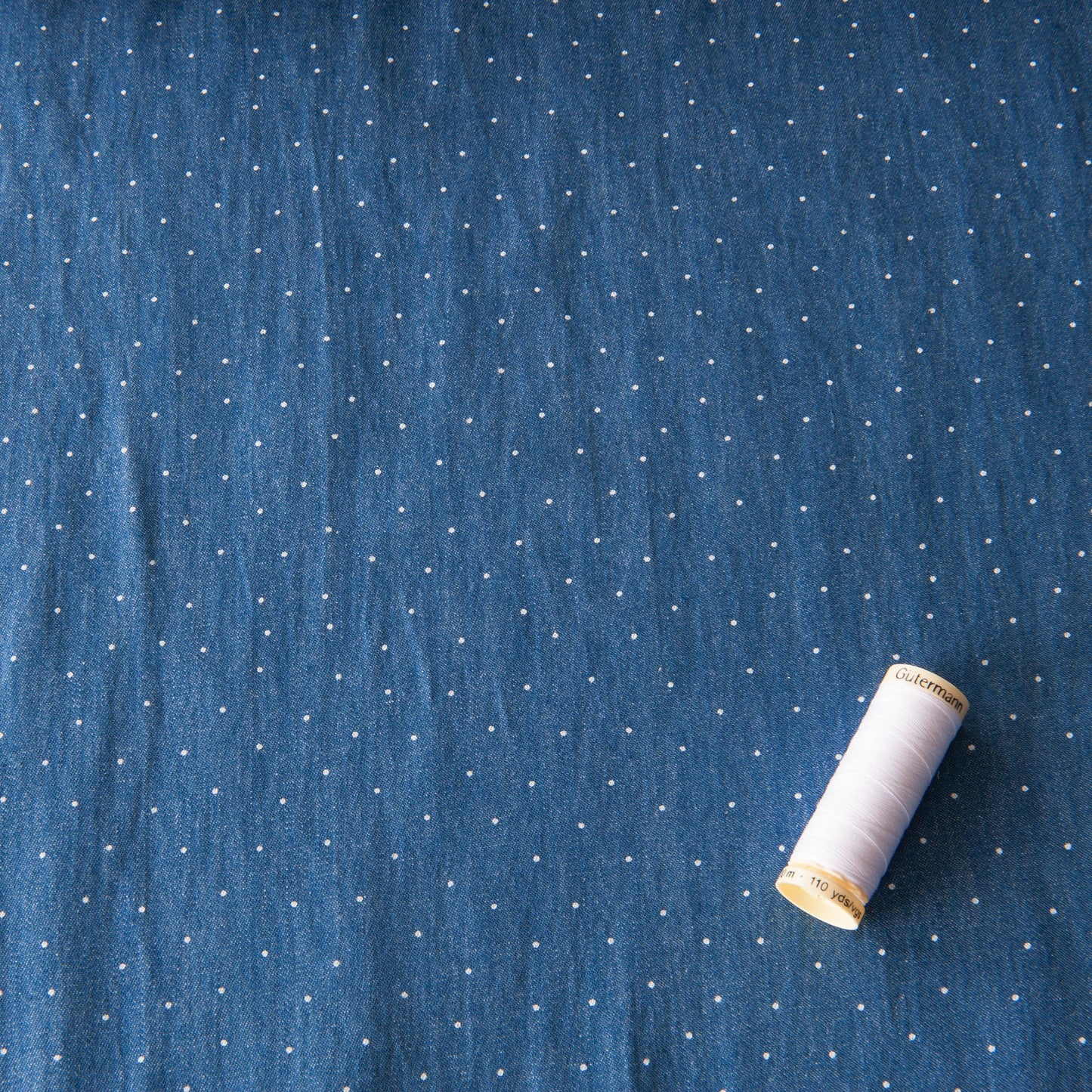 Dotty chambray dressmaking fabric with a reel of white Gutermann thread for scale