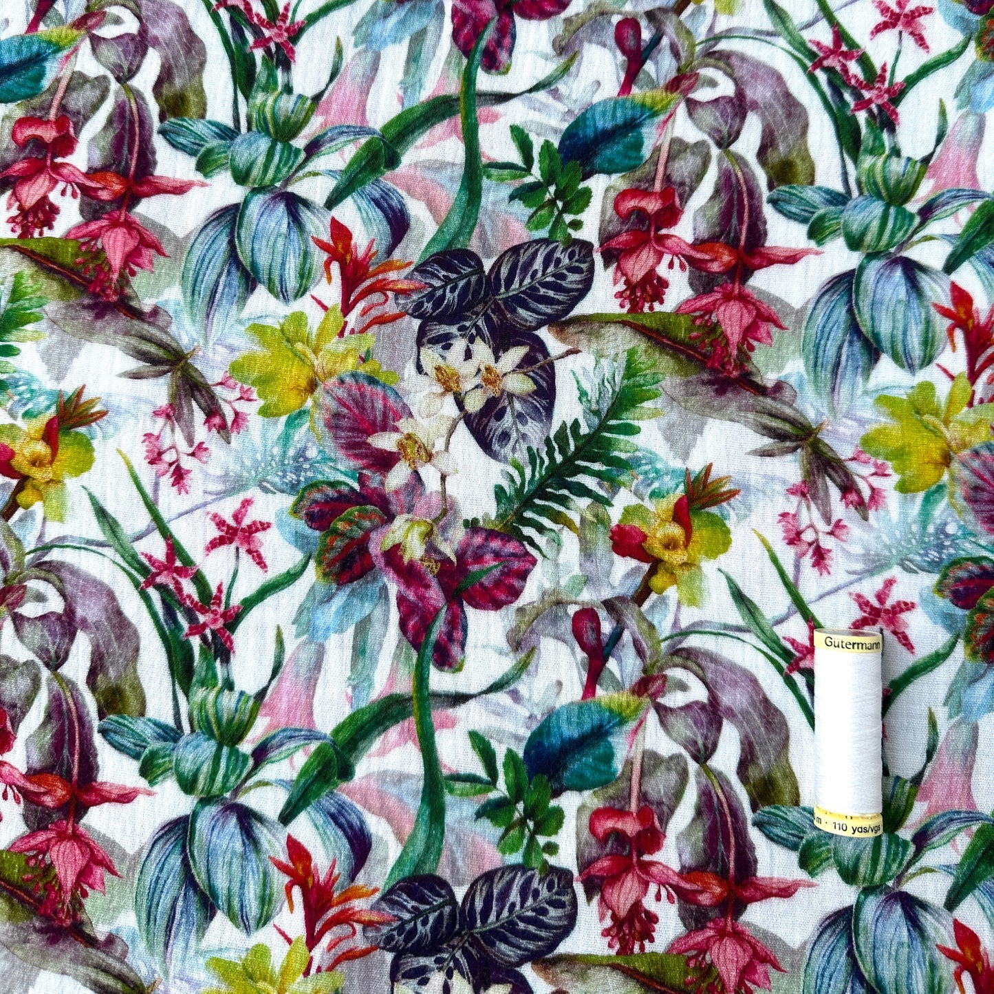 White reel of thread to show the scale of the colourful floral pattern with various flowers and leaves on a white background