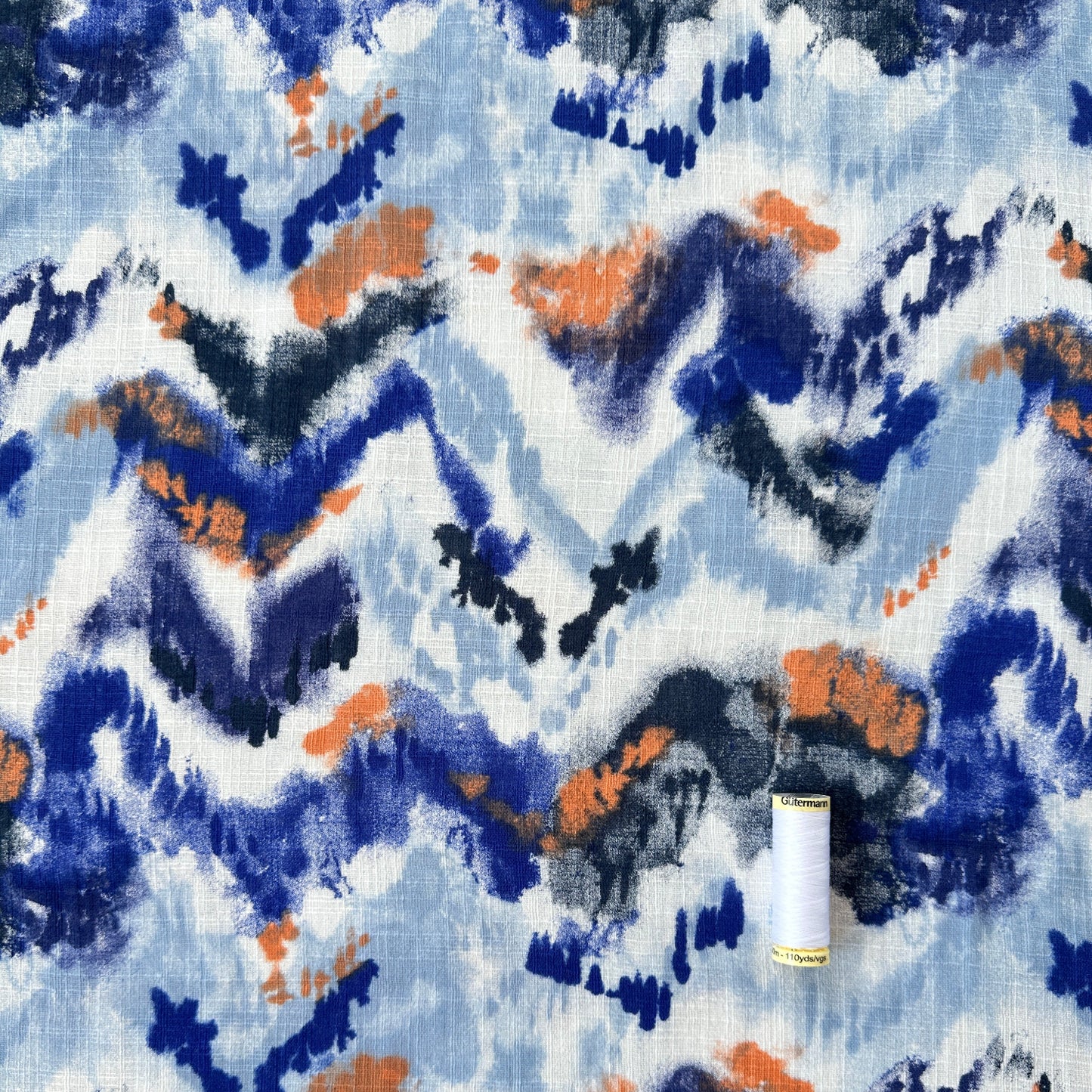 A close-up of a fabric pattern featuring an abstract, painterly design in shades of blue, navy, orange, and charcoal gray on a white background