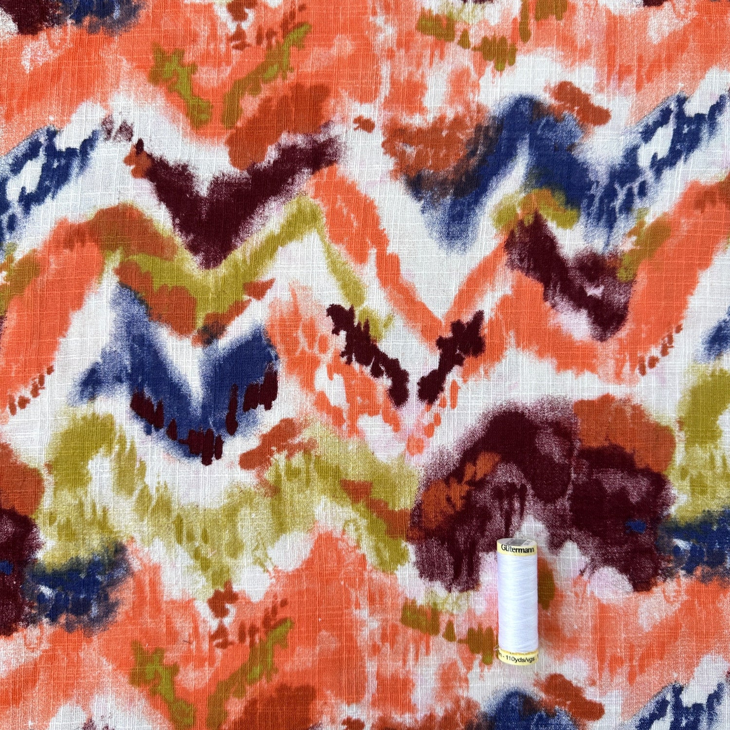 A close-up of a fabric featuring an abstract chevron design in orange, navy blue, burgundy, olive green, and white. A small white spool of thread is placed on top of the pattern as a scale or texture reference.