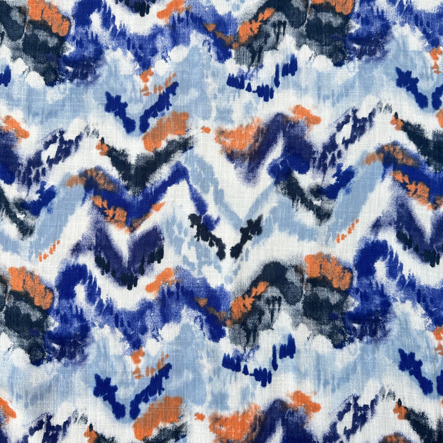 A close-up of a fabric pattern featuring an abstract, watercolor-like design with zigzag and brushstroke elements in blue, navy, orange, and white.