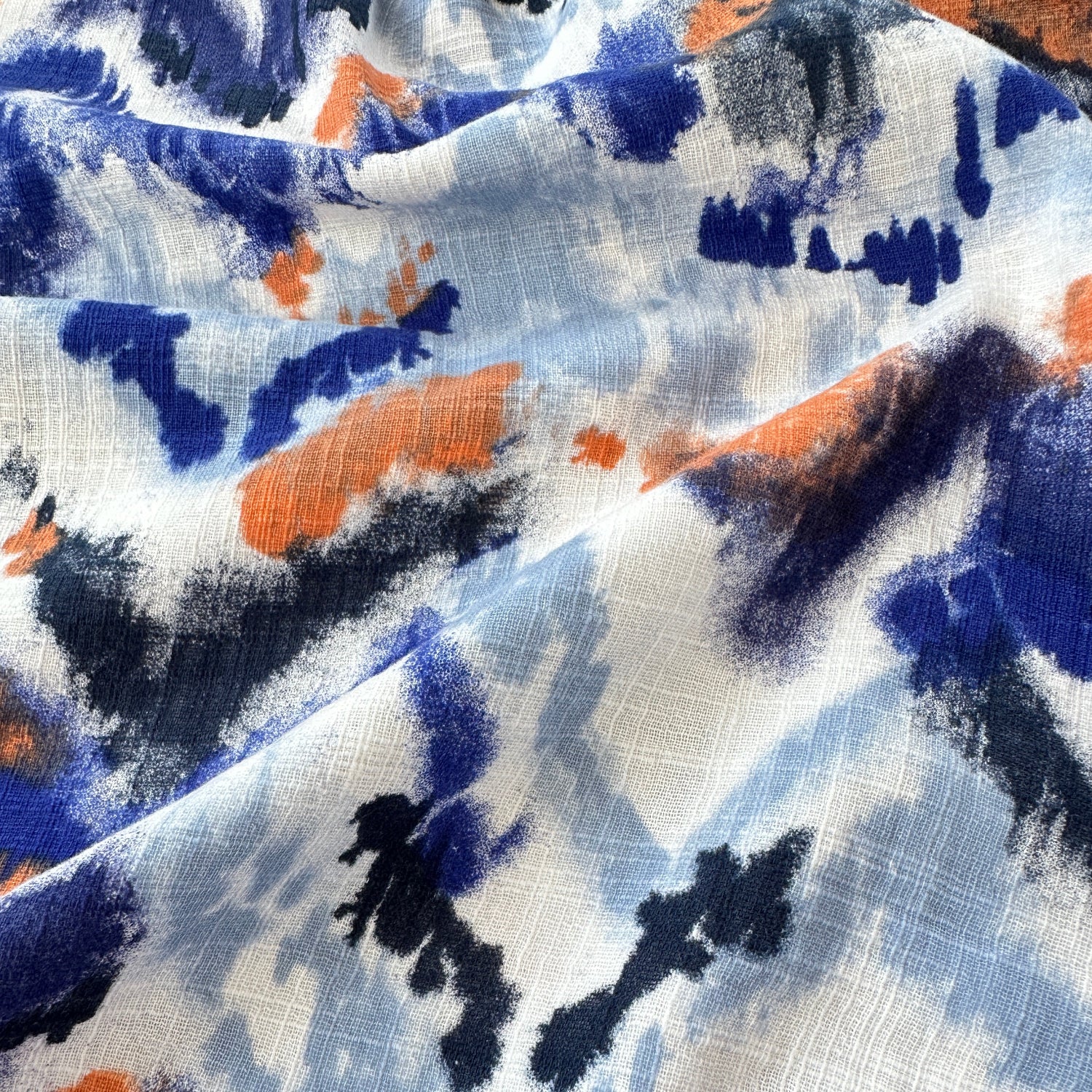 A close-up of a lightweight fabric with an abstract watercolour pattern in blue, navy, orange, and white, showing its soft texture and folds.