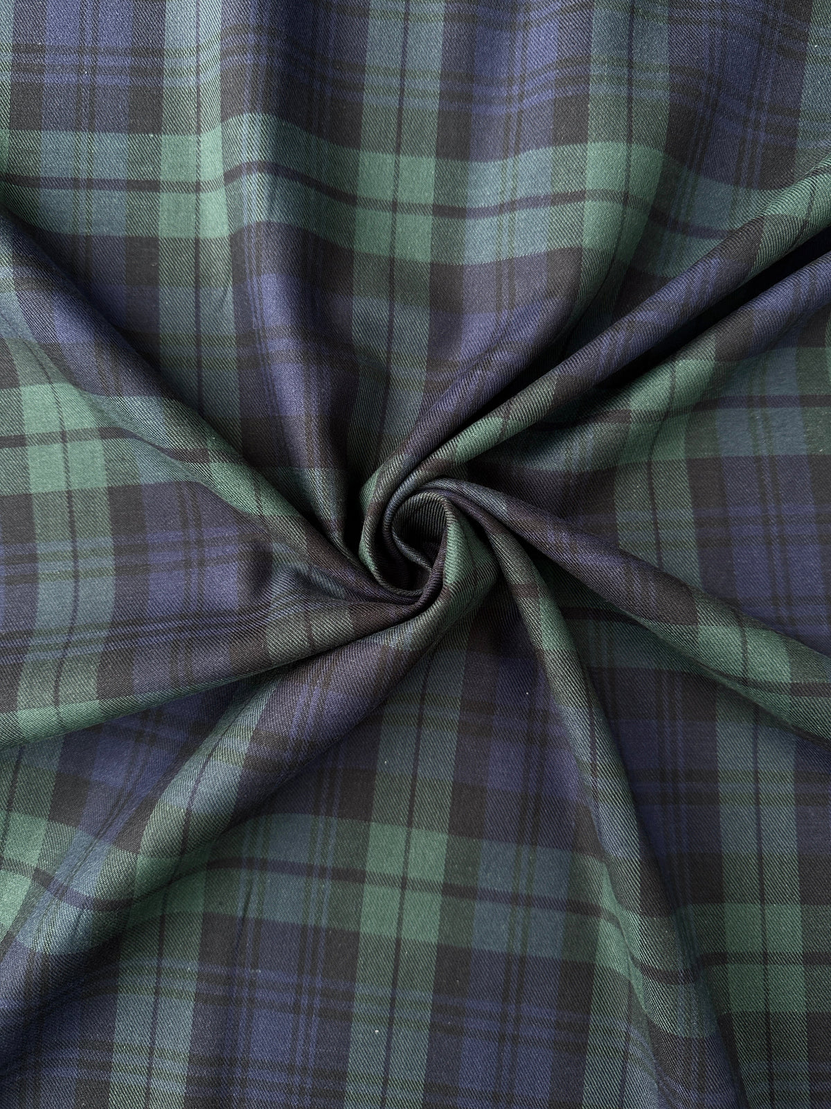 A close-up of plaid fabric arranged in a spiral drape, showing a tartan pattern in green, navy and black
