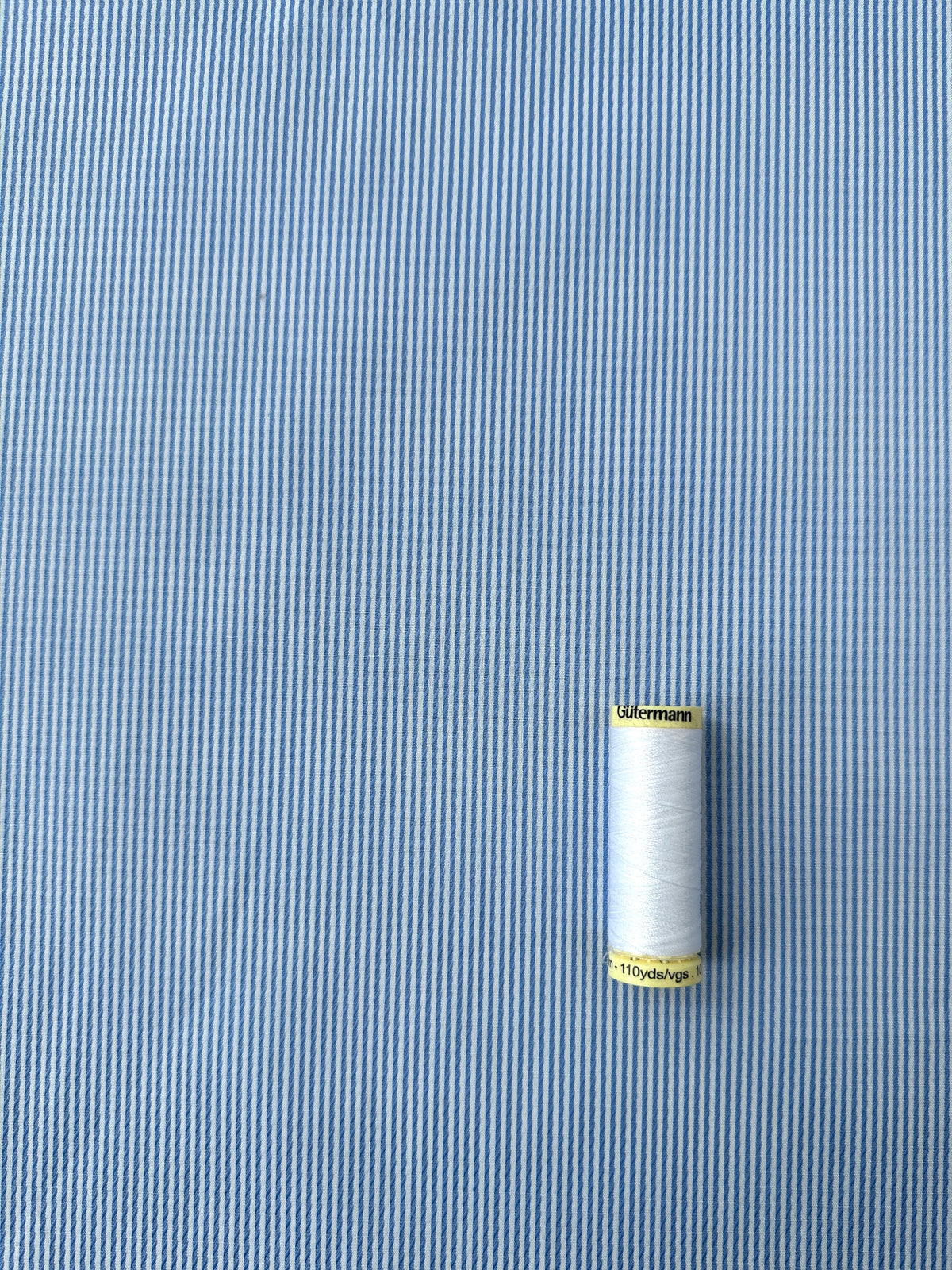 A spool of white Gutermann thread placed on a blue and white striped fabric background.