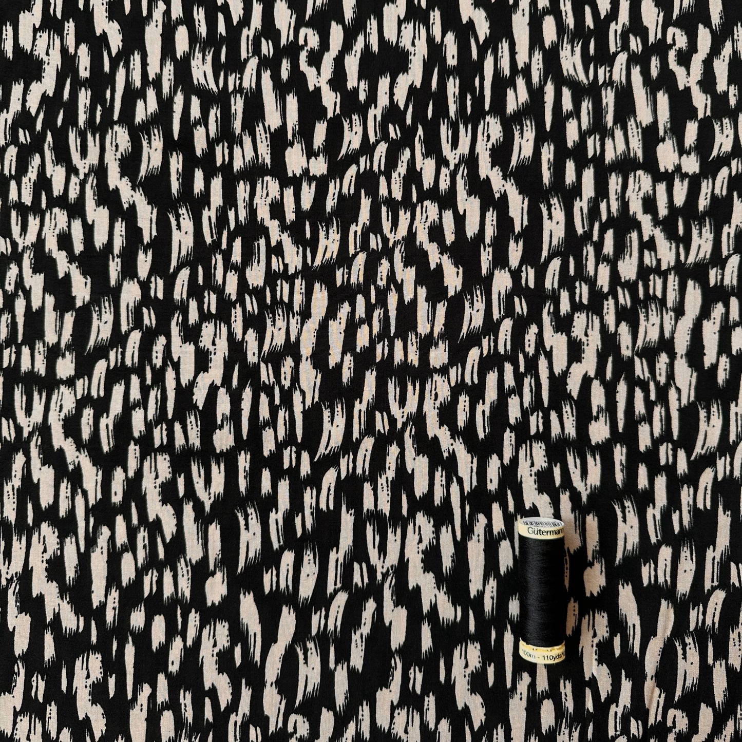 Black and fawn / beige abstract brushstroke patterned fabric shown close up, with a small black spool of thread for scale