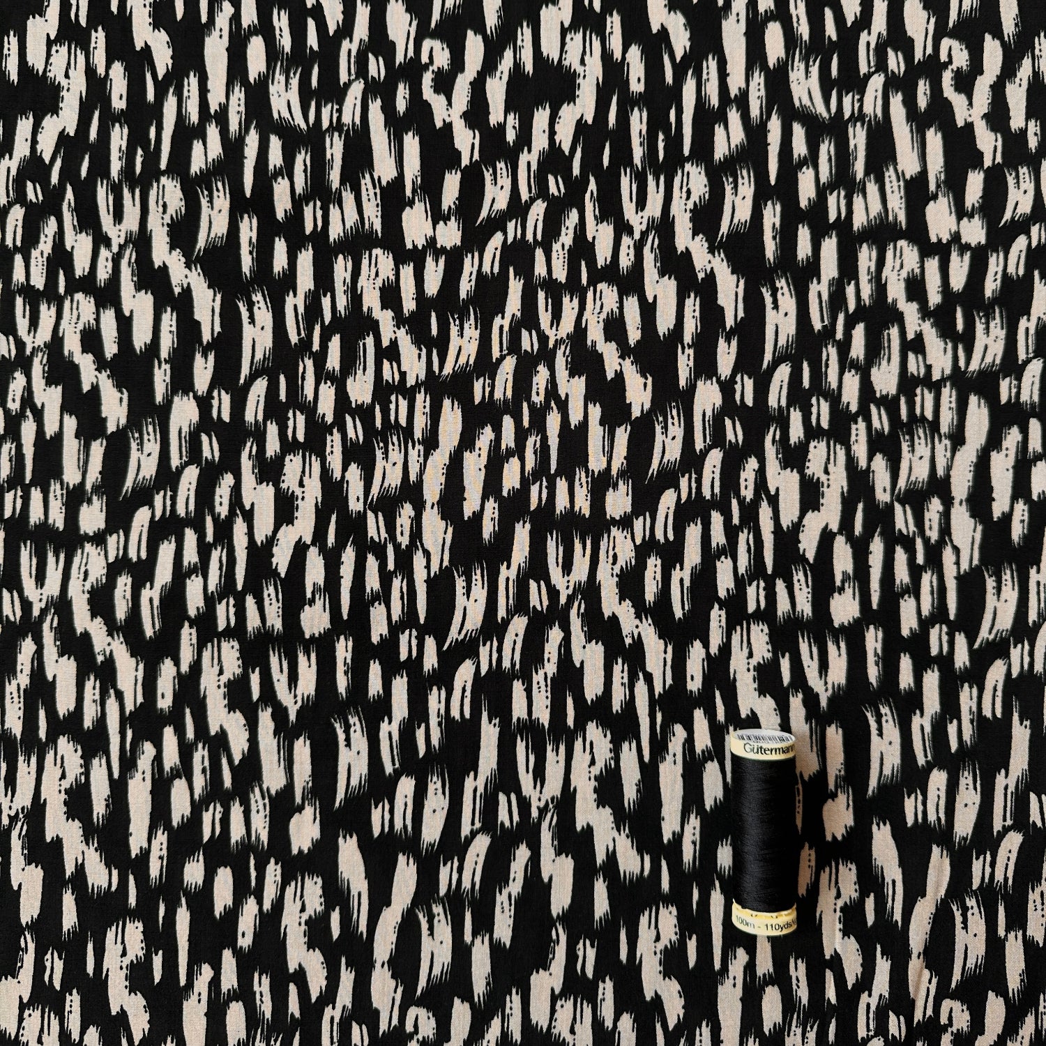 Black and fawn / beige abstract brushstroke patterned fabric shown close up, with a small black spool of thread for scale