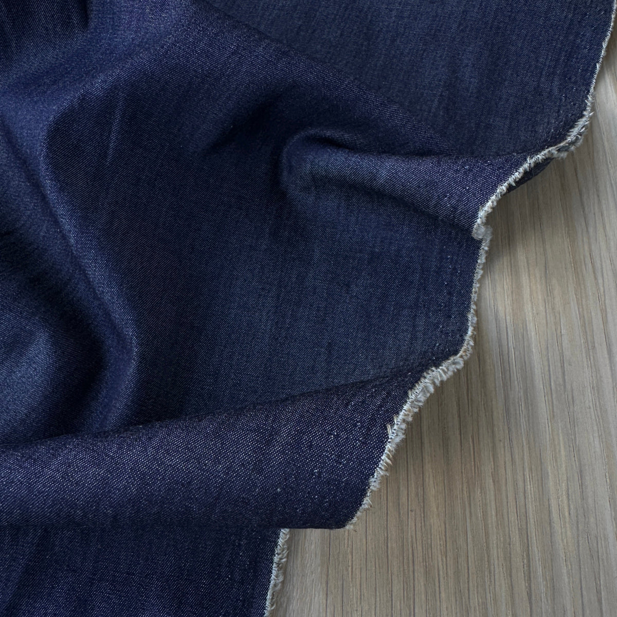 A close-up of dark navy blue fabric draped over a light wood surface, showing the material texture and selvedge