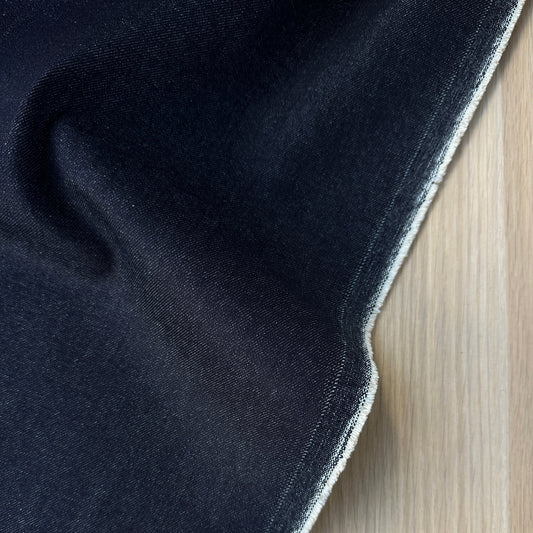 A close-up of a dark indigo denim fabric, showing the texture and selvedge edge