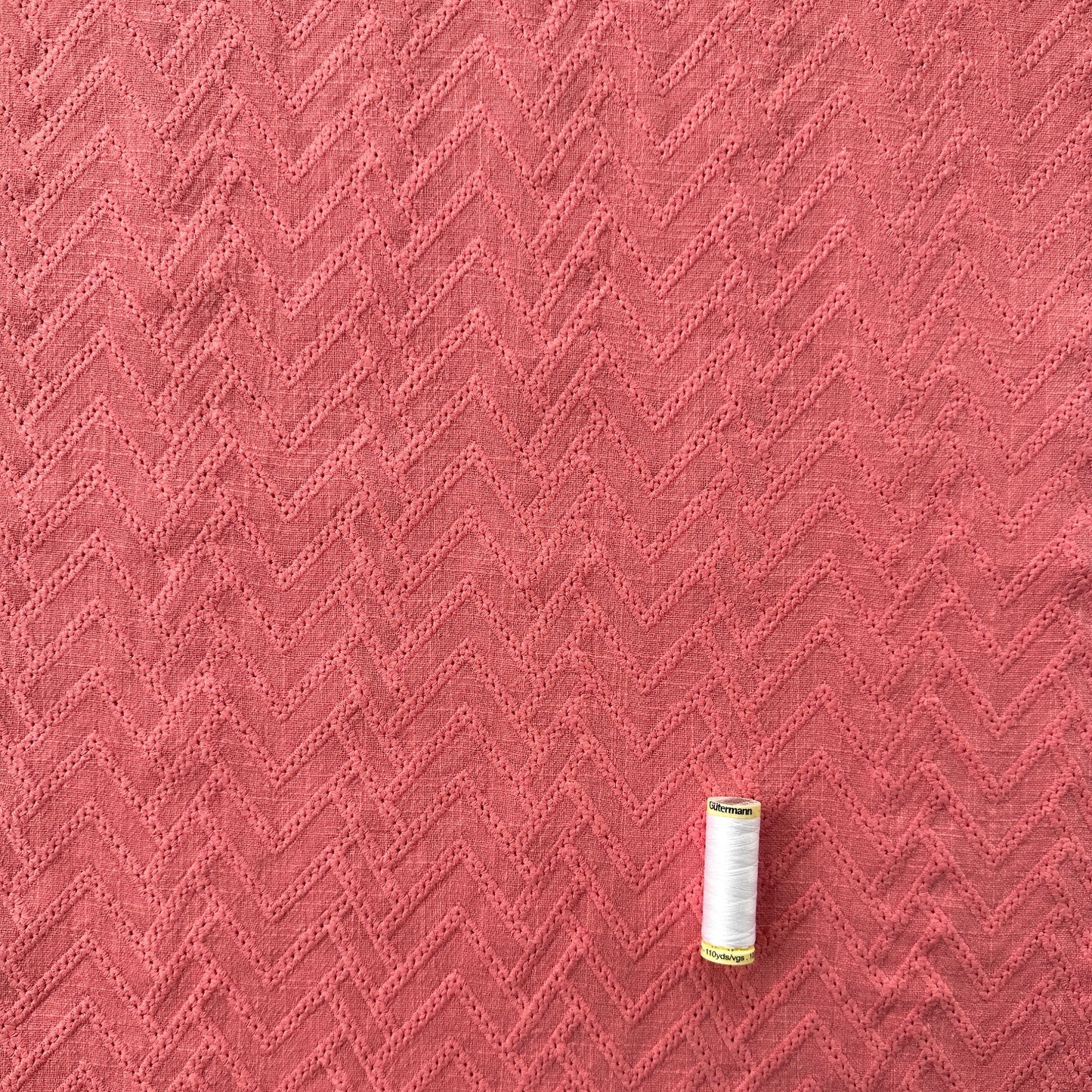 A close-up of a pink textured fabric with a woven chevron pattern, with a reel of thread for scale