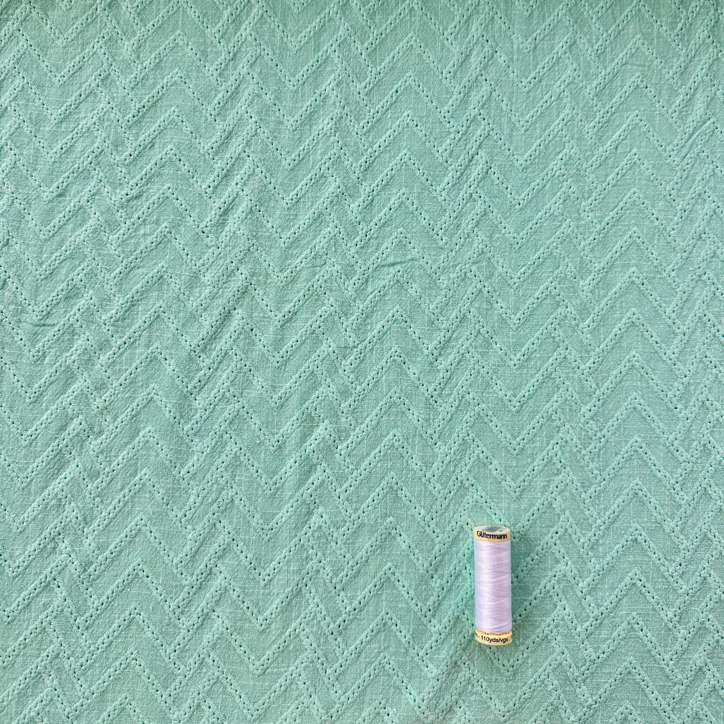 A close-up of mint green textured fabric with a subtle chevron pattern, with a small spool of thread  for scale.
