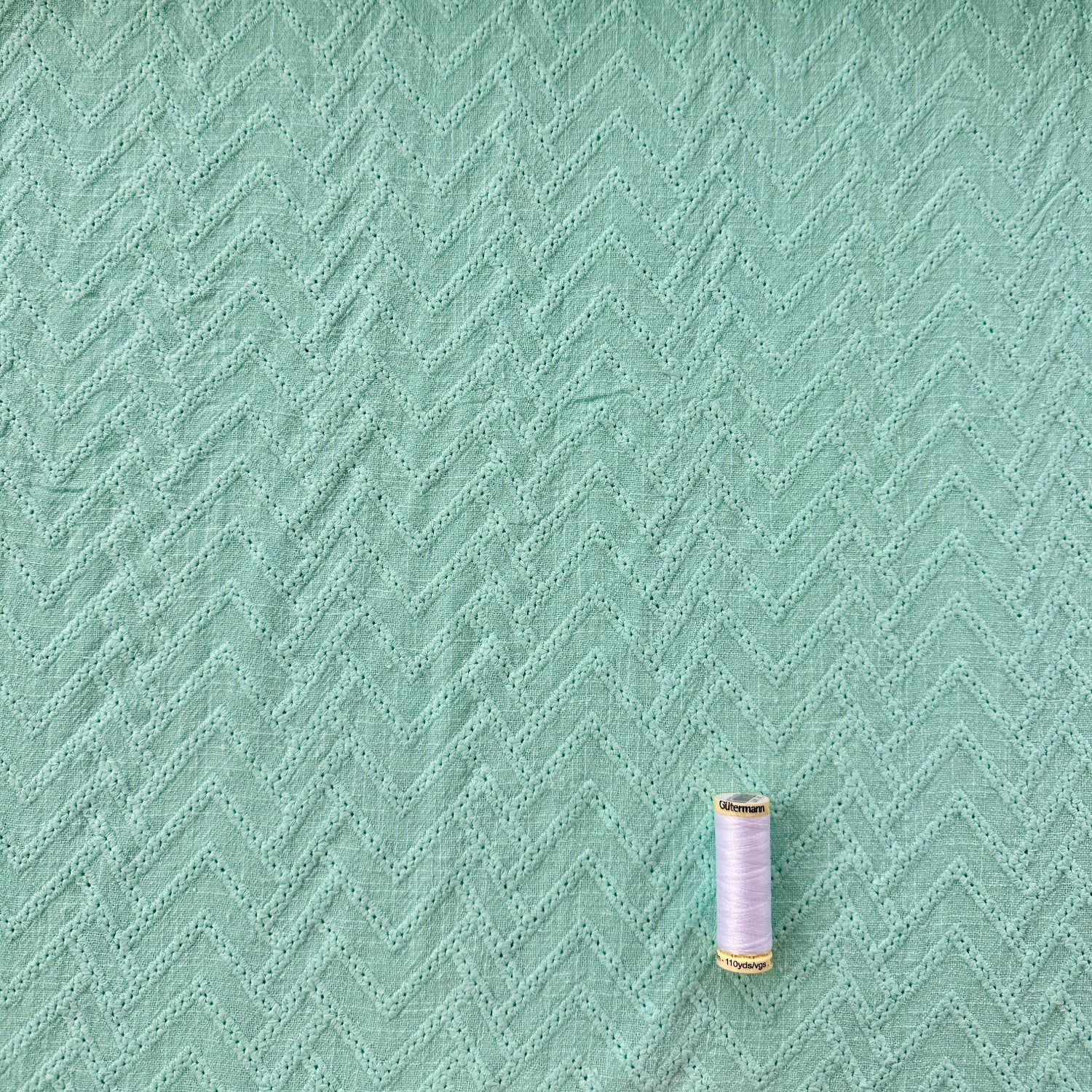 A close-up of mint green textured fabric with a subtle chevron pattern, with a small spool of thread  for scale.