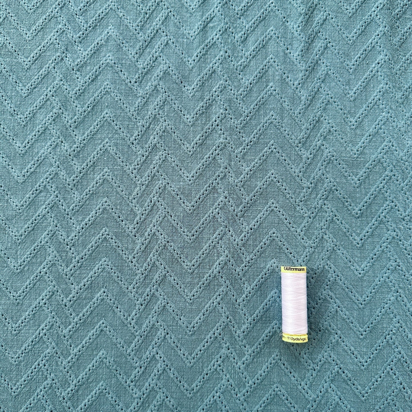 A close-up of a blue cotton fabric with embroidered zigzags across it. There is a reel of thread for scale