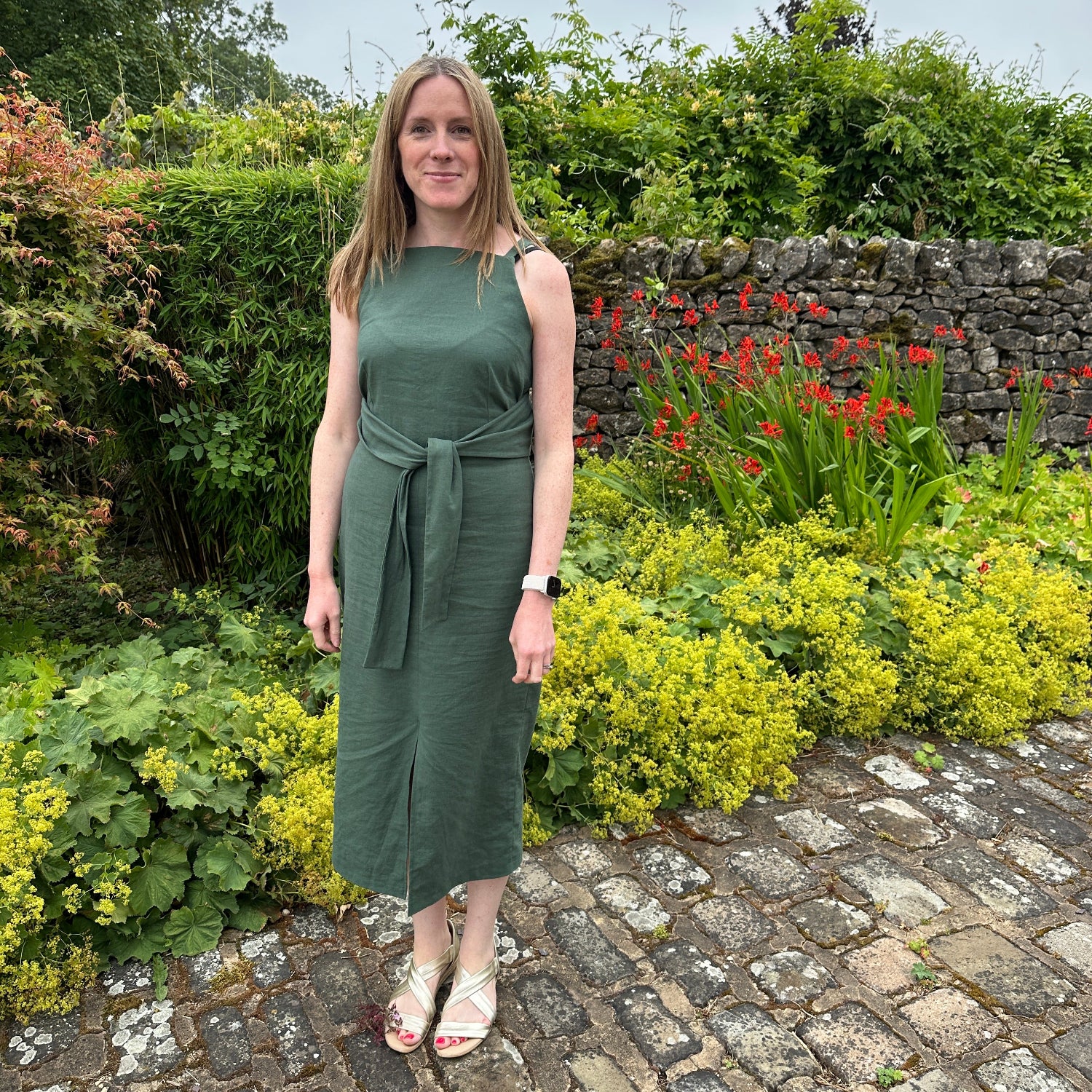 A woman wearing a sleeveless sage green dress stands outdoors on a stone path next to a garden wall with greenery and red flowers.