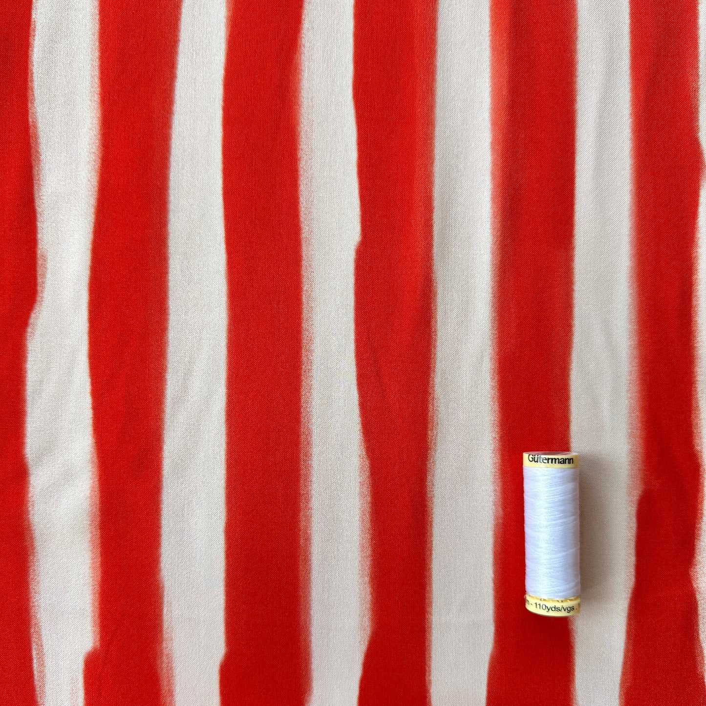 A close-up of an orangey red and ;ight peach vertically striped fabric, with a reel of thread for scale