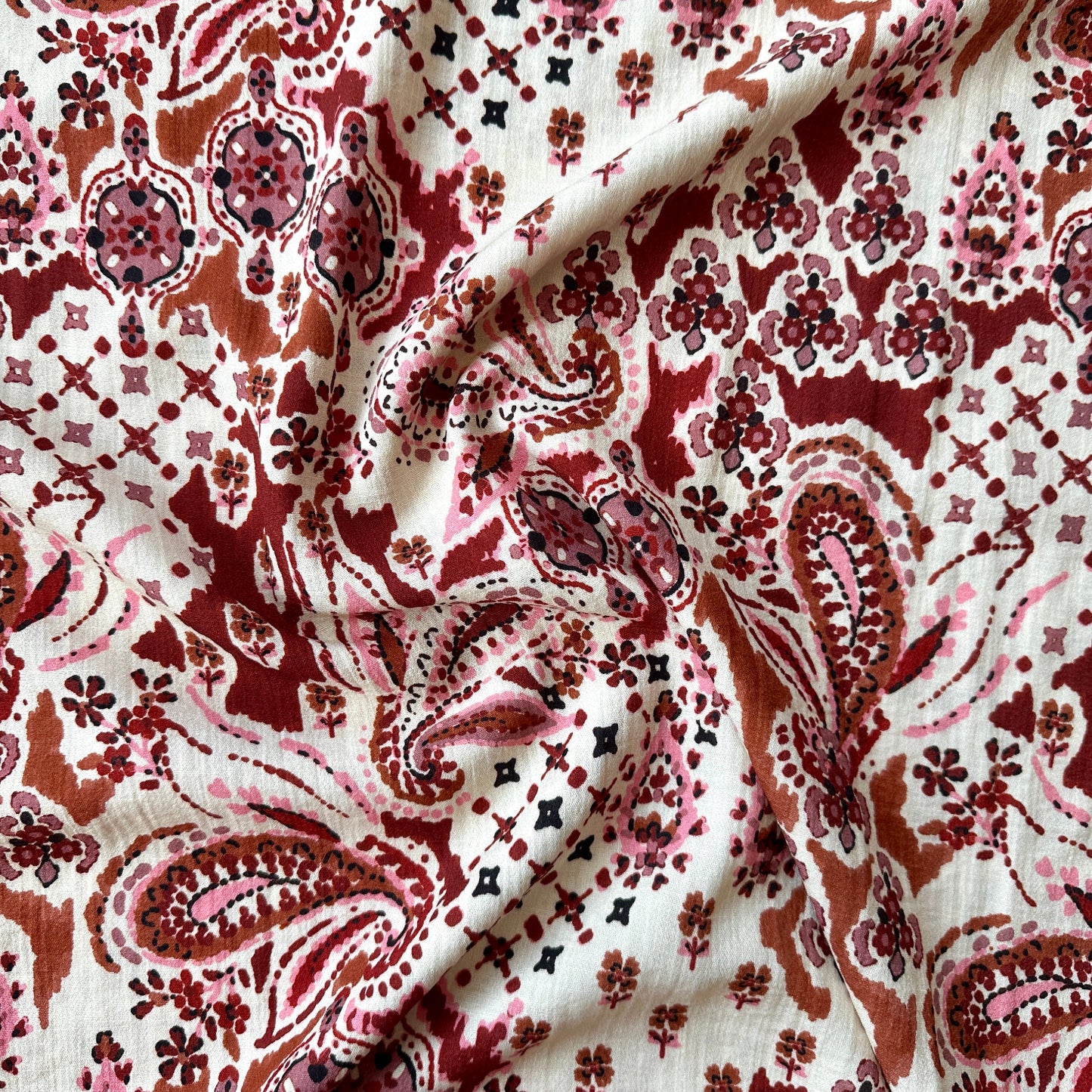 A close-up of draped fabric with an intricate paisley and floral pattern in rust red, dusty pink, and brown on a cream background