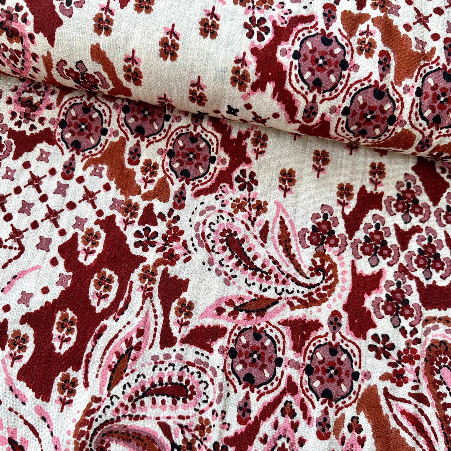 A close-up of patterned fabric with a paisley and floral print in deep red, dusty rose pink, and brown on a cream background
