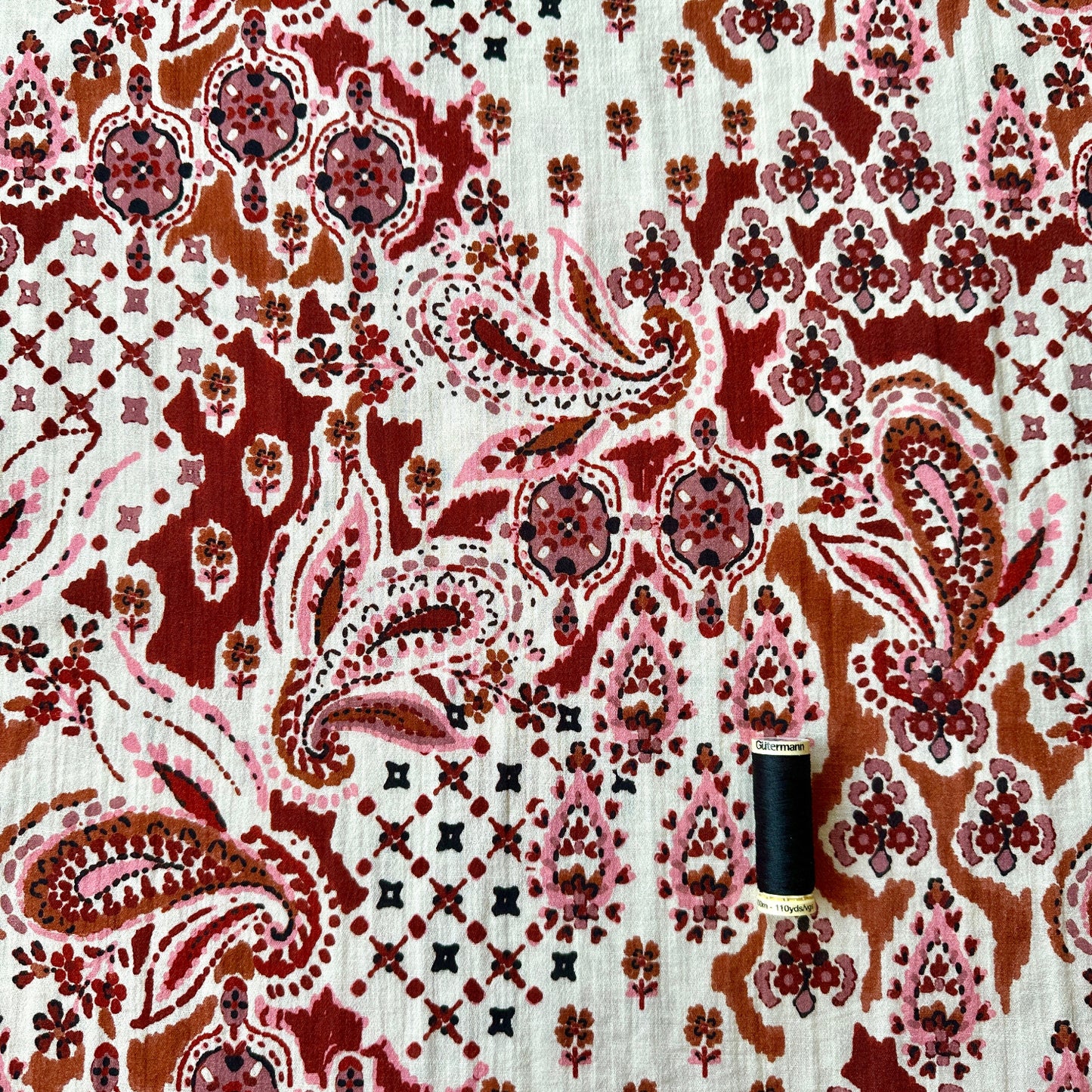 Paisley design in pinks, reds, browns on cream background with reel of black thread for scale