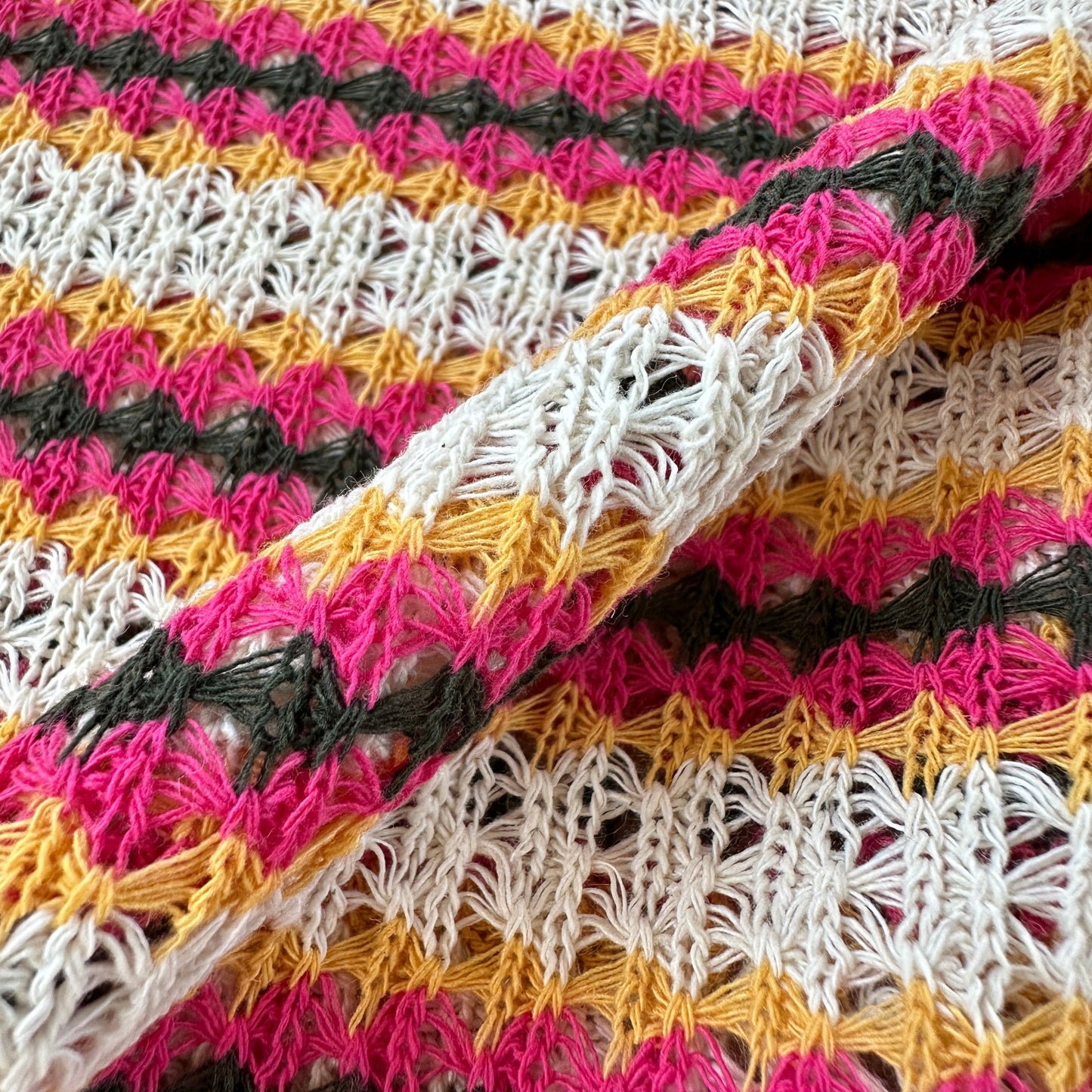 Close-up of a knitted fabric with repeating vertical stripes in ecru, pink, mustard yellow, and grey, showing the textured crochet pattern and folded fabric.