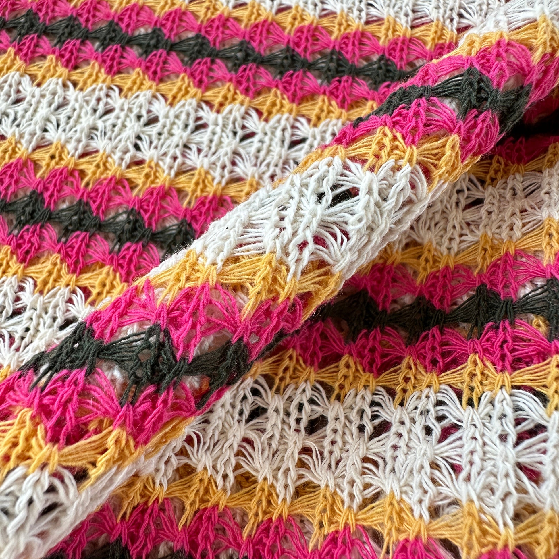 Close-up of a knitted fabric with repeating vertical stripes in ecru, pink, mustard yellow, and grey, showing the textured crochet pattern and folded fabric.