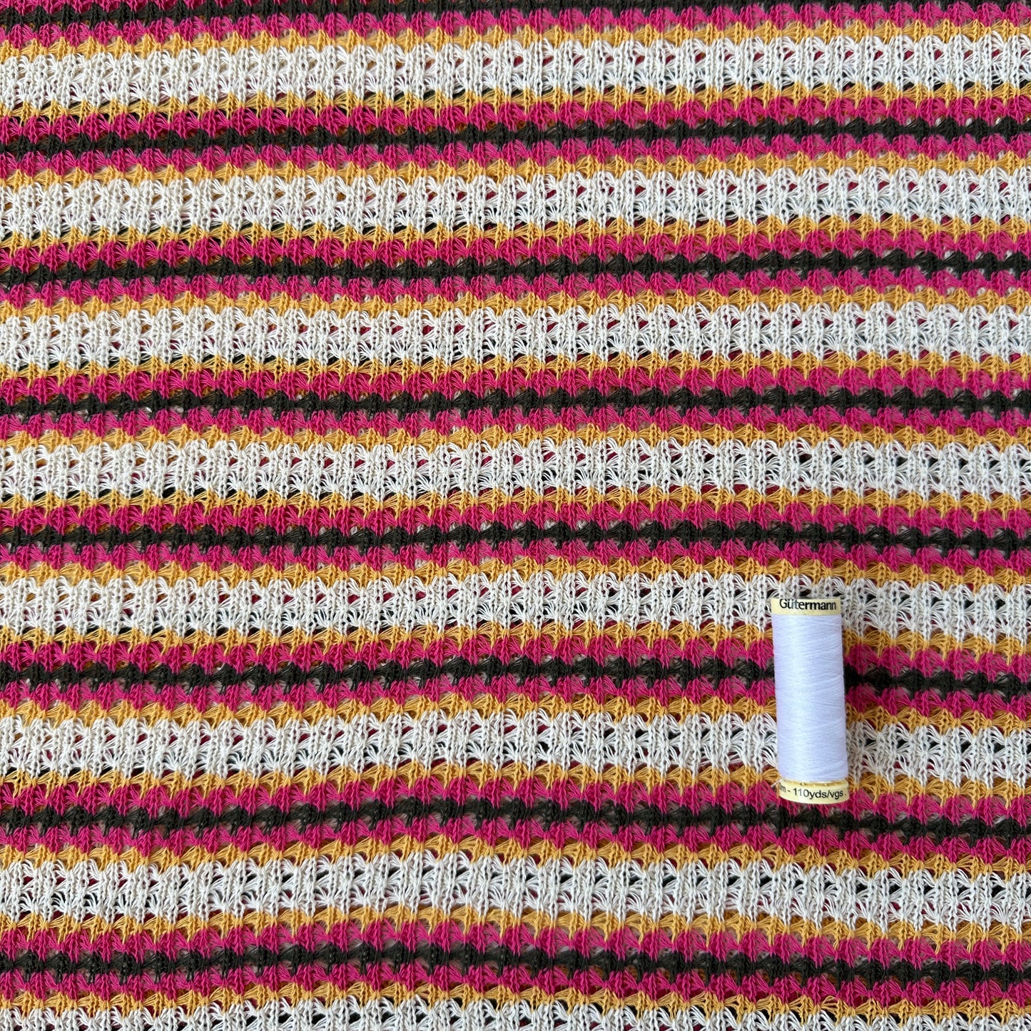 A close-up of a crocheted fabric with vertical stripes in ecru, pink, grey and yellow, with a small spool of thread resting on top.