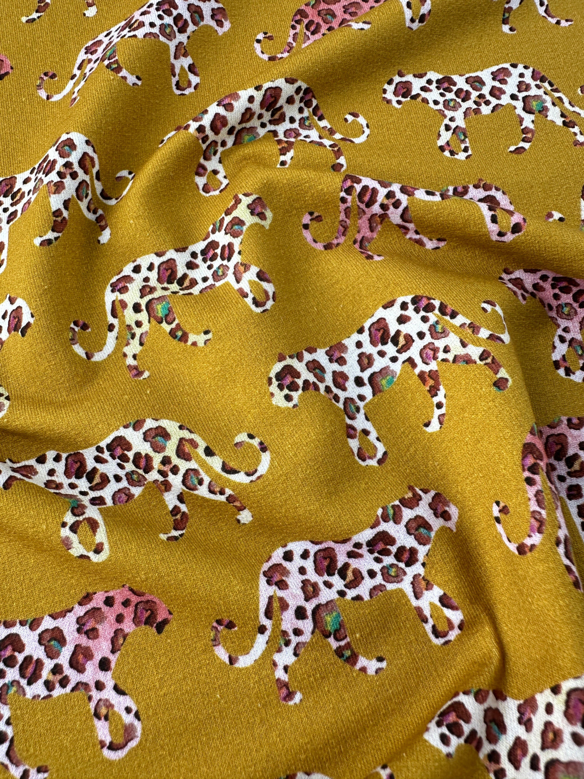 A close-up of draped fabric featuring a repeating pattern of walking leopard silhouettes in white, pink, and brown on a mustard yellow background.