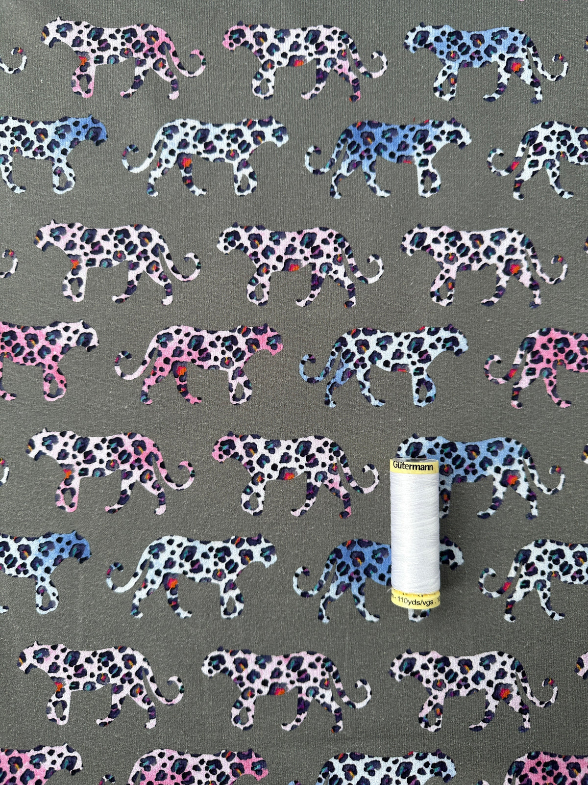 A close-up of fabric featuring a repeating pattern of walking spotted animals in pink, blue, black, and white on a grey background, with a spool of thread for scale