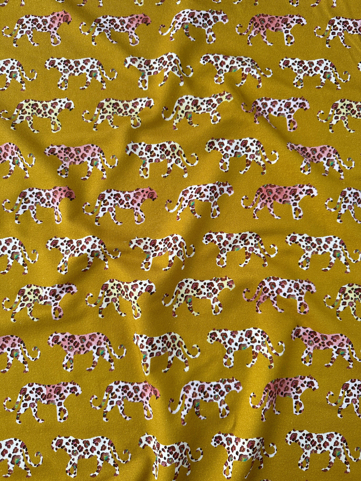 A close-up of a mustard yellow fabric with a repeating pattern of leopards walking in white, pink, and brown.