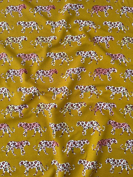 A close-up of a mustard yellow fabric with a repeating pattern of leopards walking in white, pink, and brown.
