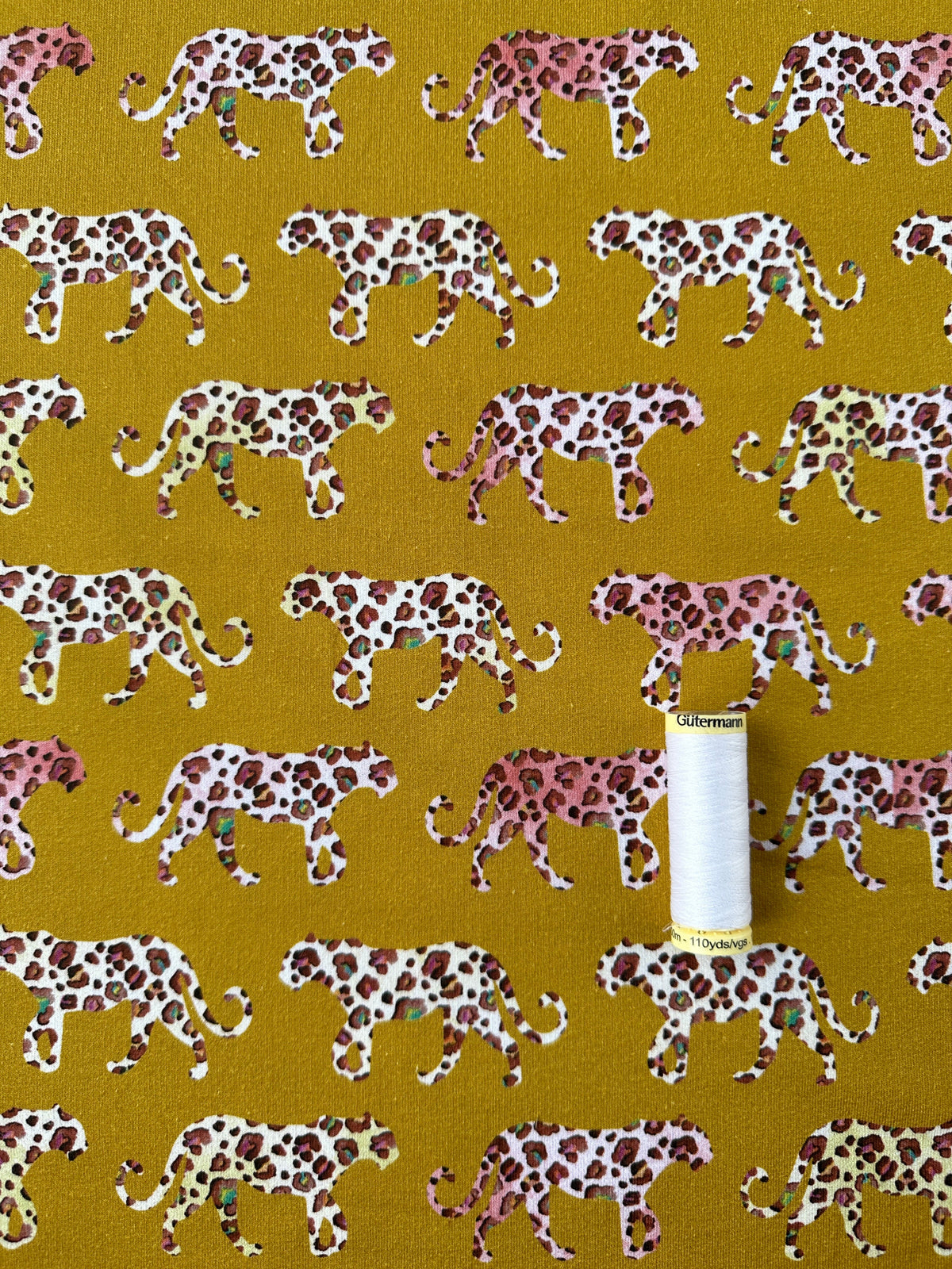 A close-up of patterned fabric featuring repeated leopard silhouettes in pink, brown, and cream on a mustard yellow background, with a white spool of thread placed on the surface.