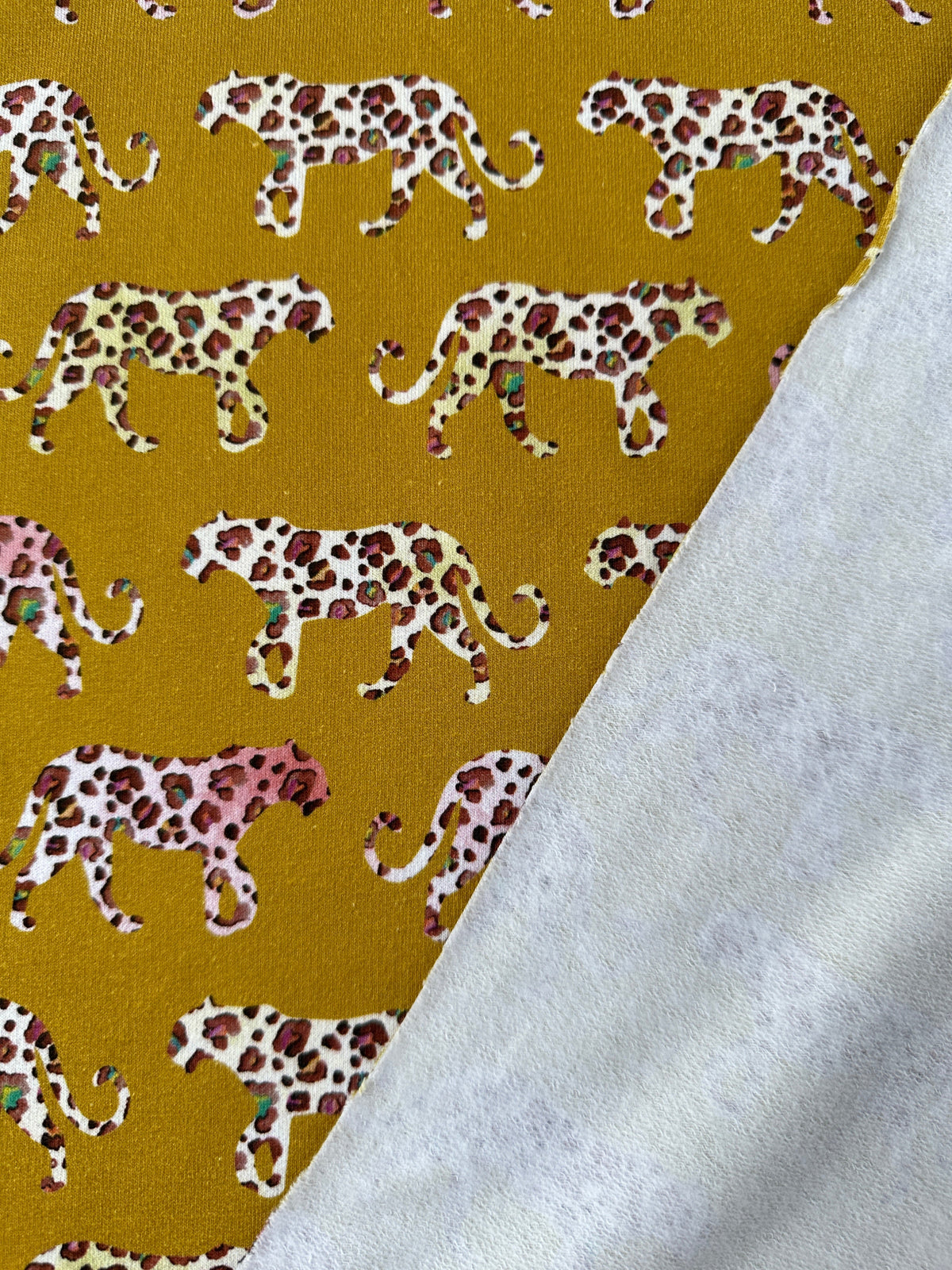 A close-up of patterned fabric with walking leopard silhouettes in white, pink, and brown on a mustard yellow background, with a the back of the fabric showing