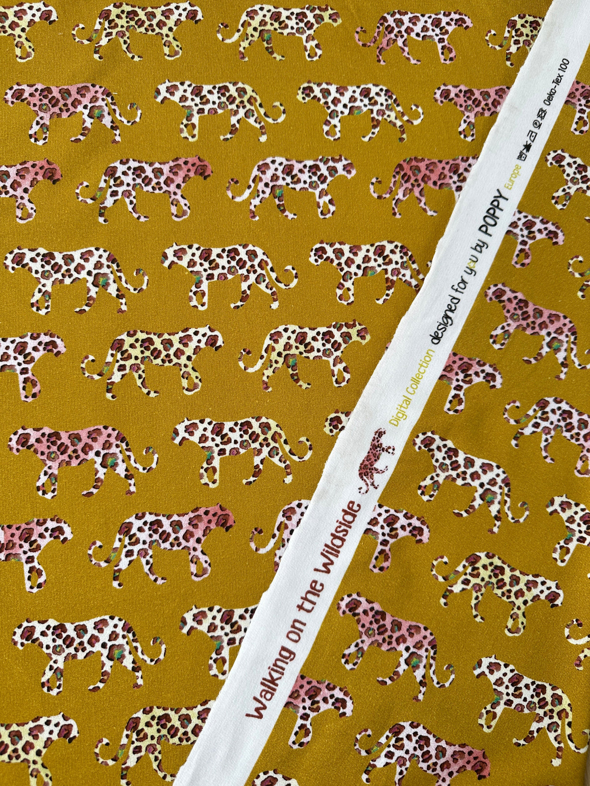 A close-up of a fabric featuring walking leopards in pink, brown, and white on a mustard yellow background, with the white Poppy Europe selvedge across the middle